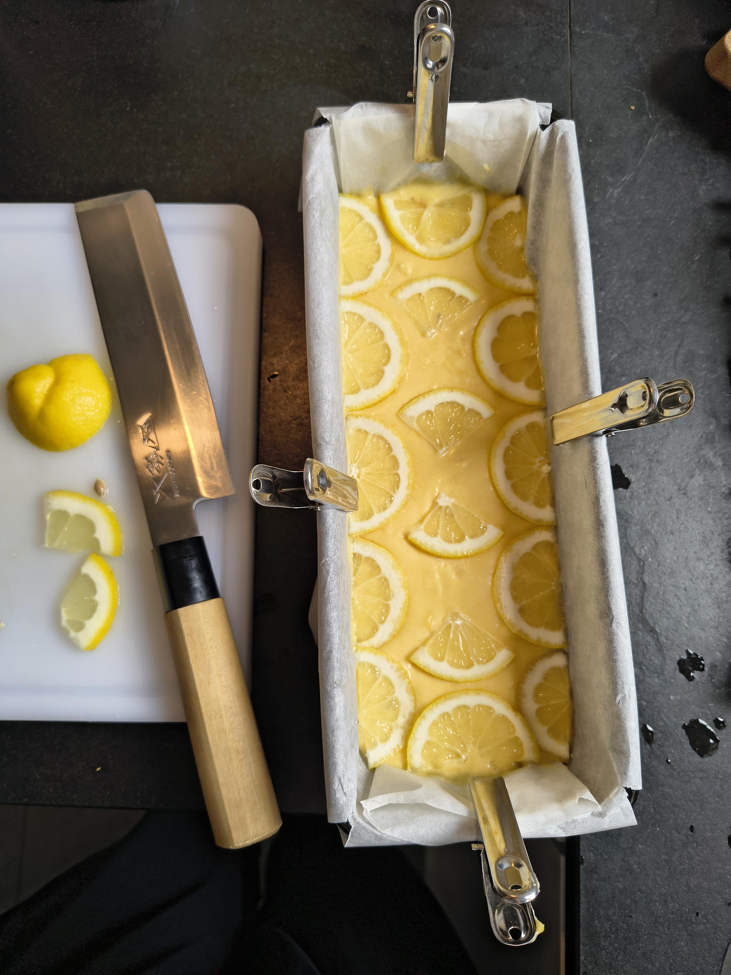 Un bizcocho de limón y curcuma a punto de ser metido al horno. Es una masa muy amarilla (por la curcuma) y lleva rodajas finitas de limón decorando por encima. Al lado está la tabla de cortar, el cuchillo y restos de limón.